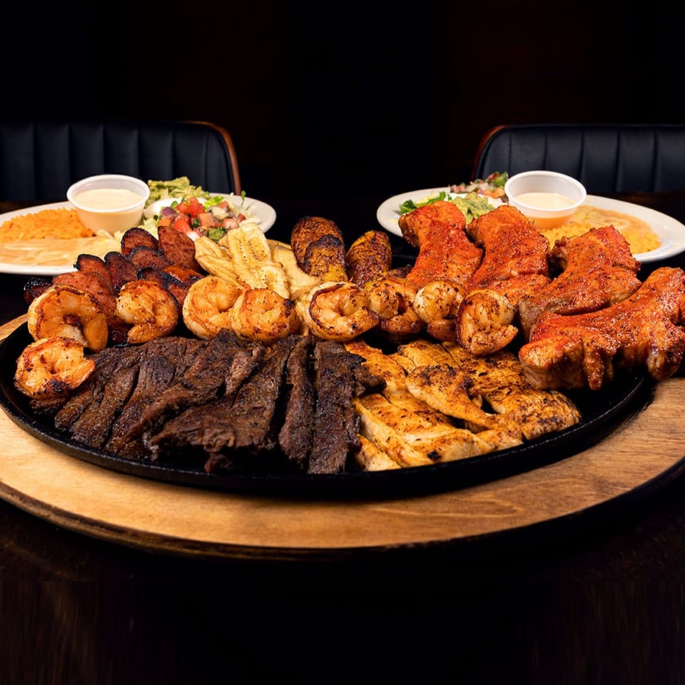 Large sizzling platter of grilled steak, chicken, shrimp, chorizo, and plantains with sides.