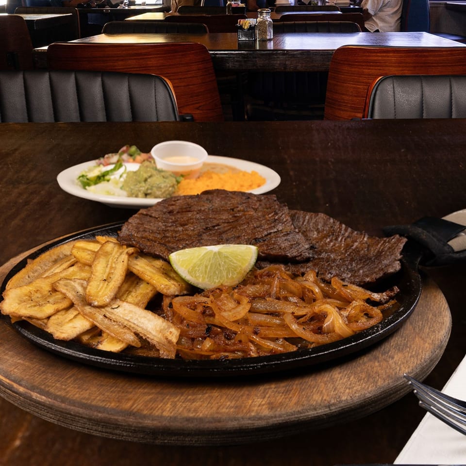Sizzling steak fajitas with plantains, caramelized onions, and lime, with a restaurant interior in the background.