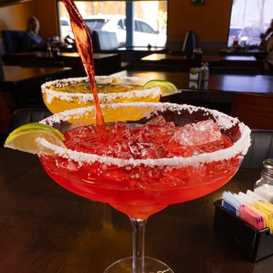 Red margarita with ice and lime being poured, with a yellow margarita in the background.