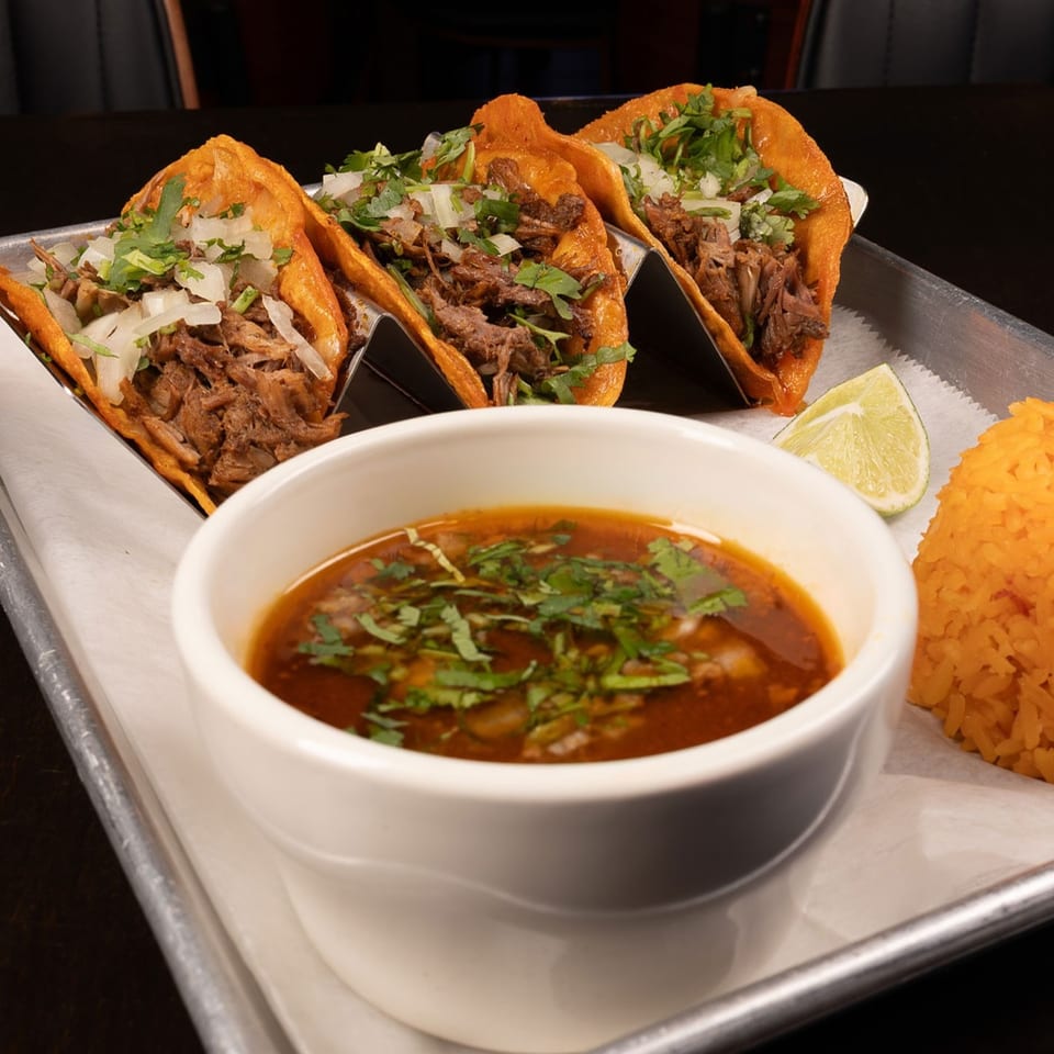 Three birria tacos with cilantro and onions, a side of consommé, and orange rice.