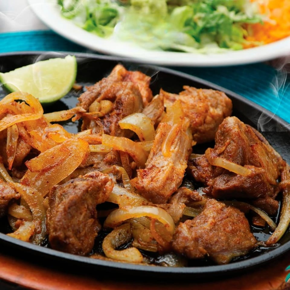 Sizzling Cantina Del Mar fajita meat and onions with a lime wedge, and a salad in the background.