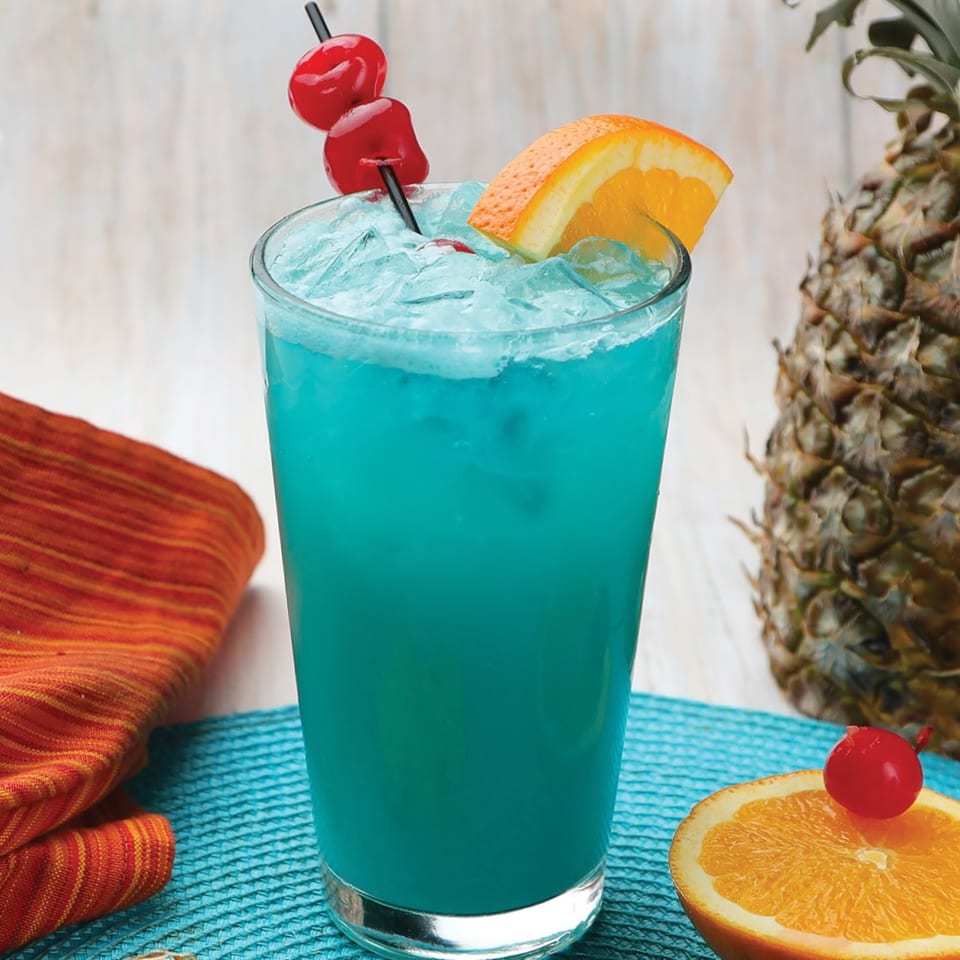 Vibrant blue cocktail with ice, garnished with cherries and an orange slice, next to a pineapple.