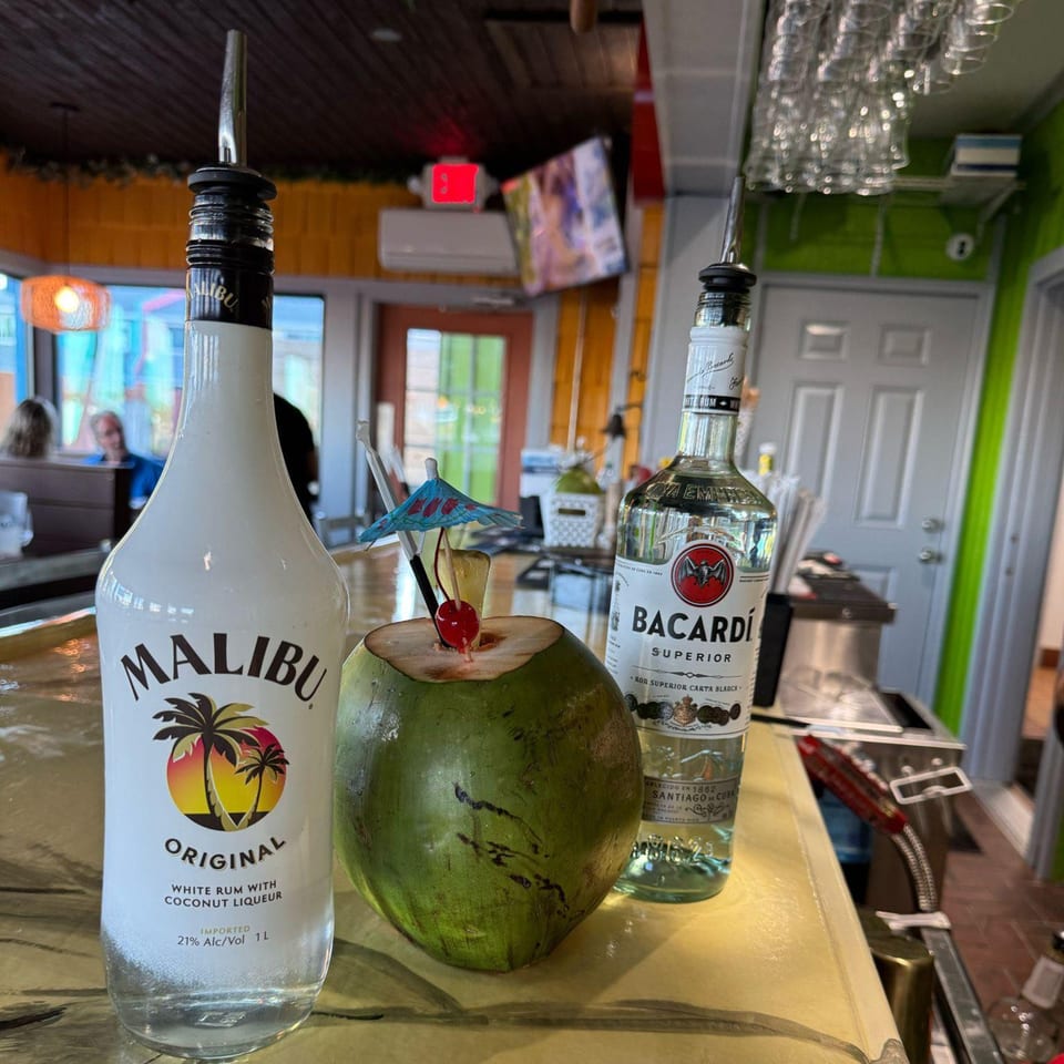 A fresh coconut drink with a cherry and umbrella, flanked by Malibu and Bacardi rum bottles on a bar.