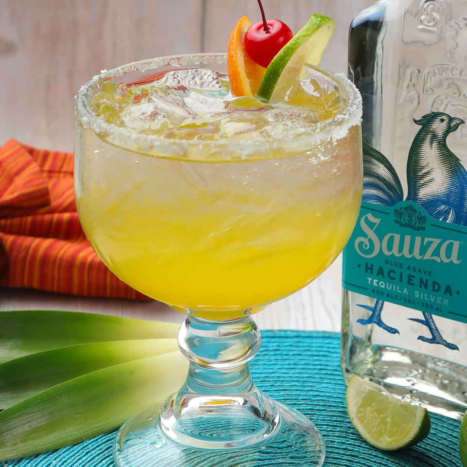 A vibrant yellow margarita in a salt-rimmed glass, garnished with orange, lime, and a cherry.